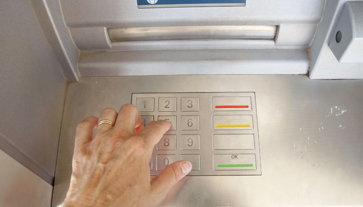 Na zdjęciu widzimy rękę osoby dotykającą klawiatury numerycznej bankomatu. Klawiatura składa się z przycisków z cyframi od 1 do 9 rozmieszczonymi w trzech rzędach po trzy oraz przyciskiem 0 poniżej. Po prawej stronie klawiatury znajdują się dodatkowe przyciski z kolorowymi paskami: czerwonym, żółtym i zielonym, prawdopodobnie służące do potwierdzania operacji, anulowania lub wyjścia. Klawiatura jest metalowa i umieszczona na przednim panelu bankomatu o szarej barwie. W tle widoczna jest metalowa struktura bankomatu, a samo zdjęcie jest wykonane w naturalnym świetle.
