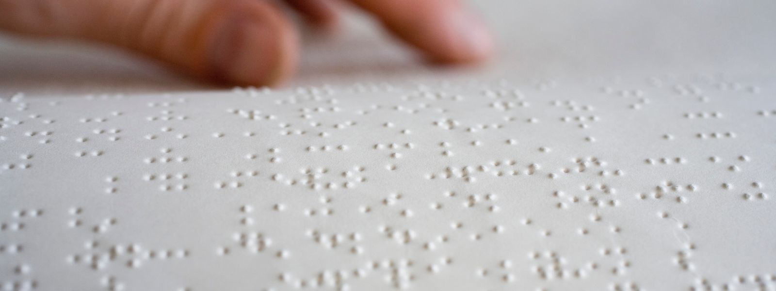 Na zdjęciu widoczna jest zbliżona perspektywa białej kartki z wypukłym pismem Braille'a. Punkty są regularnie rozmieszczone w rzędach, tworząc znaki alfabetyczne. W tle nieostro widzimy palec osoby, który dotyka tekstu, sugerując proces czytania przez dotyk. Jasne oświetlenie podkreśla wypukłość kropek, co pozwala na lepszą percepcję wzrokową tekstu w piśmie Braille'a.
