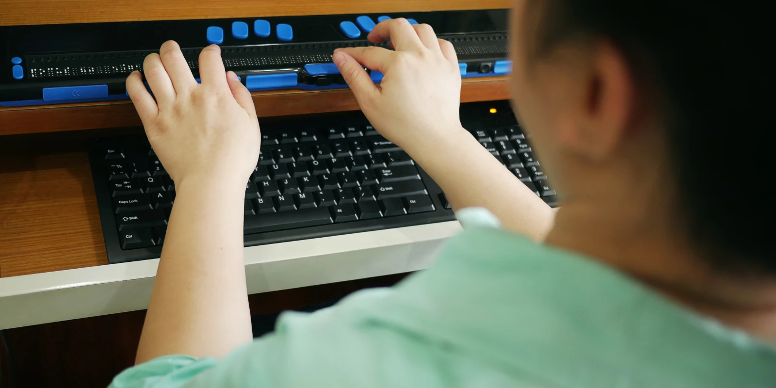 Na zdjęciu widzimy osobę z niepełnosprawnością, która korzysta z linijki Braille'a umieszczonej na biurku, nad klawiaturą komputera. Linijka Braille'a to urządzenie, które umożliwia osobom niewidomym lub niedowidzącym czytanie tekstu wyświetlanego na ekranie komputera poprzez wypukłe kropki tworzące alfabet Braille'a. Osoba ma położone dłonie na linijce, co sugeruje, że czyta lub orientuje się w tekście. Obok linijki Braille'a znajduje się standardowa klawiatura komputera. Całość wygląda na środowisko pracy dostosowane do potrzeb osób z dysfunkcją wzroku.